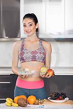 Young fit woman in the kitchen holding fruits and macarons