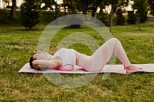 young fit woman in pink sportswear lies with back on pilates ball to stretch and relax back muscles in park in summer