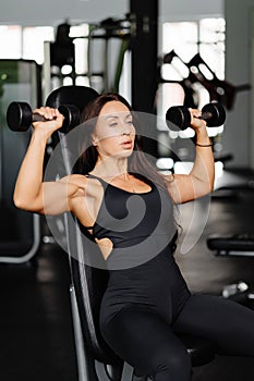 Young fit woman practicing dumbbell press while sitting on a bench. Slim female in sportswear exercises with weights in a gym