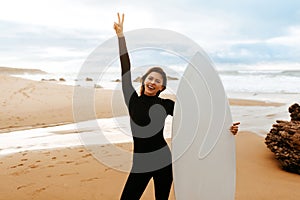 Young fit woman standing with surfboard on beach by seaside, smiling at camera and gesturing peace sign