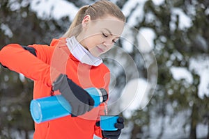 Young fit woman in warm fitness clothes pours hot tea into mug from metal thermos, after workout outdoors on sports ground on