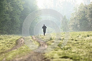Young fitness man running at dawn on wild forest road into haze.