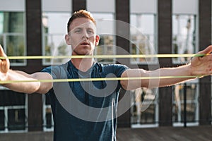 Young fitness man in sportswear doing exercises with elastic hand resistance band on building background