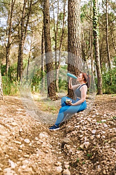 young fitness woman sitting in a forest resting after training. sporty girl drinking water from a bottle to refresh