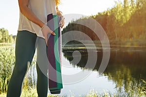 Young fitness woman standing by the lake with yoga mats, close-up. Yogi woman practice meditation at beautiful sunset.