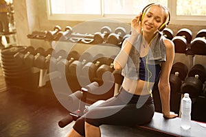 young fitness woman with towel listening music with headphones with water bottle relaxing sitting on dumbbell rack in gym . sport