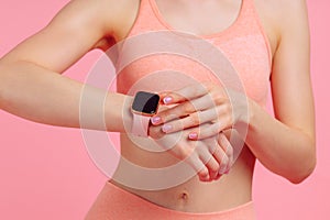 Young fitness woman wearing headphones in sportswear resting after workout using smartwatch on pink background