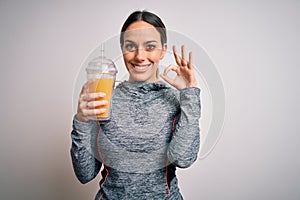 Young fitness woman wearing sport workout clothes drinking fresh orange juice doing ok sign with fingers, excellent symbol