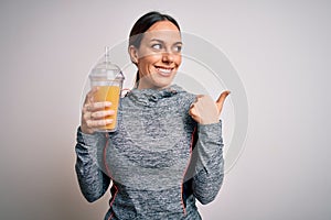 Young fitness woman wearing sport workout clothes drinking fresh orange juice pointing and showing with thumb up to the side with