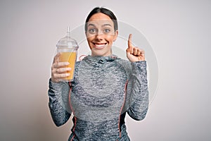 Young fitness woman wearing sport workout clothes drinking fresh orange juice surprised with an idea or question pointing finger