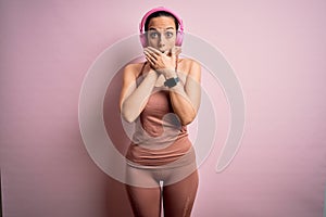 Young fitness woman wearing sport workout clothes wearing headphones listening to music shocked covering mouth with hands for