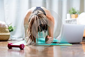 Young fitness woman working out and using laptop and dumbbells at home in living room, doing yoga or pilates exercise on