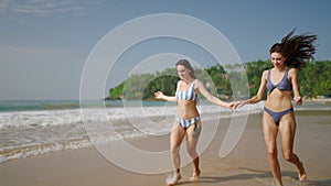 Young fitted women in swimwear holding hands, running along sea water edge. Smiling pretty girls run, make water
