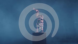 Young flexible blonde circus acrobat posing in studio in costume. Doing equilibre balance handstand on a cube.