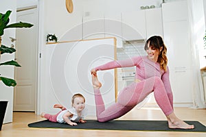 Young flexible mother engaged in stretching workout with her baby at home