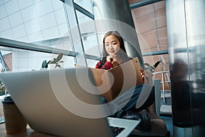 Young focused asian female IT employee holding open copybook and watching laptop