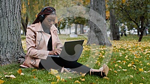 Young focused female student freelancer sitting in autumn park near tree working on laptop typing message checks email