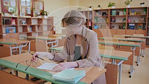 A young focused teacher female sits in a classroom and checks the students' homework by taking notebooks from a pile