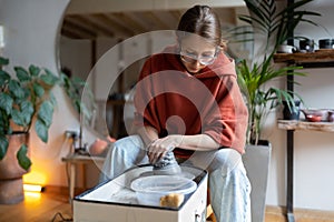 Young focused woman local ceramic artist working with clay on wheel in cozy ceramics studio