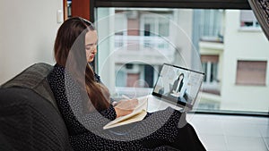 Young focused woman sitting on sofa at home, making notes during distant chat with psychologist