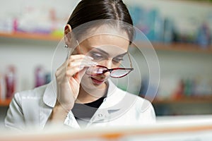 A young focussed brown-haired girl with glasses, wearing a lab coat, stands in a pharmacy by holding her glasses and