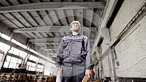 Young foreman standing in the building plant with welding in the background. Worker is holding construction plan and