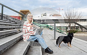 Young freelance expat woman sitting on the stairs of office building drinking coffee and working on laptop computer with her dog