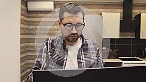 Young freelance in glasses sitting at the kitchen and typing on laptop