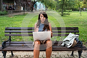 Young freelance modern hipster woman working online in the park sitting on the bench having video call on web cam on computer. E-
