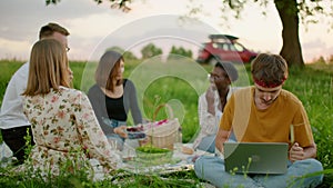 Young freelancer Man works on Laptop while friends have Picnic Party outdoors