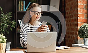 Young freelancer   works on a laptop and searches for information in your smartphone in her home office