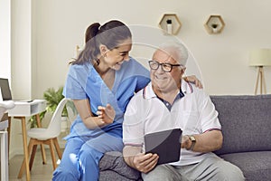 Young friendly female caregiver and senior man using digital tablet together in nursing home.