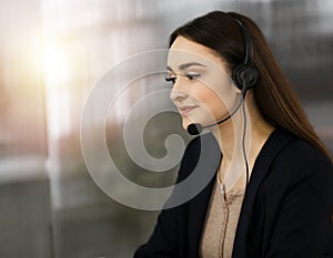 Young friendly girl in headsets is talking to a firm`s client, while sitting at the desk in sunny office. Call center