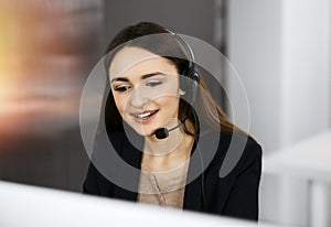 Young friendly girl in headsets is talking to a firm`s client, while sitting at the desk in sunny office. Call center