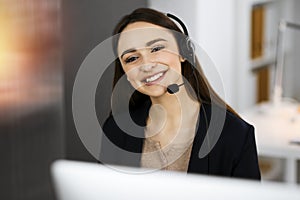 Young friendly girl in headsets is talking to a firm's client, while sitting at the desk in sunny office. Call center