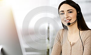 Young friendly girl in headsets is talking to a firm& x27;s client, while sitting at the desk in sunny office. Call center