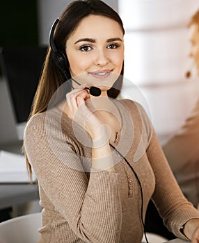 Young friendly girl in headsets is talking to a firm& x27;s client, while sitting at the desk in sunny office. Call center