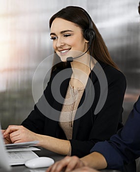 Young friendly girl in headsets is talking to a firm& x27;s client, while sitting at the desk in sunny office. Call center
