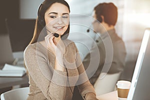 Young friendly girl in headsets is talking to a firm's client, while sitting at the desk in sunny office. Call center