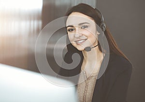 Young friendly girl in headsets is talking to a firm& x27;s client, while sitting at the desk in sunny office. Call center