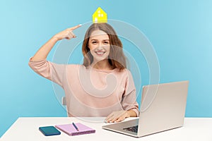 Young friendly happy woman, real estate agent sitting at workplace with laptop and pointing at small paper house