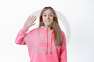 Young friendly woman saying hello, waving hand and smiling, say hi, greet you, make goodbye gesture, standing over white