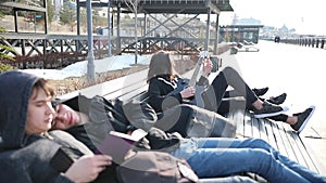 Young friends chilling on a bench - two guys reading a book - a man tuning his guitar and his girlfriend watching him