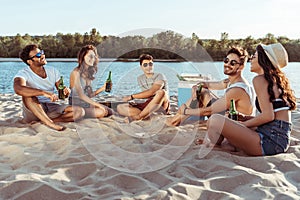 Young friends drinking beer while relaxing on sandy beach at riverside