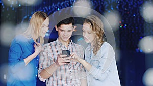 Young friends - man and two women in a nightclub use smartphone, together watching something interesting on display.