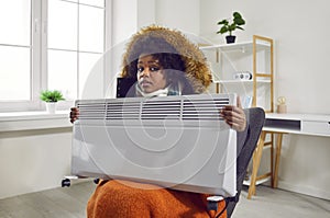 Young frozen woman sitting in a winter coat on a chair at home and holding electric heater.