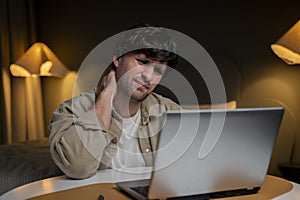 Young frustrated freelancer is experiencing severe neck pain. He is in his home office, sitting at his modern workplace