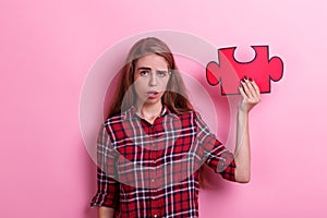 Young frustrated girl, holds a big red puzzle and grimaces. On a pink background.