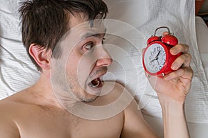 Young frustrated and stressed man is late. He is waking up, looking at alarm clock.