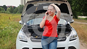 Young frustrated woman leaning on the broken car and calling service for help
